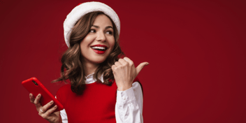 Smiling woman wearing a Santa hat, holding a smart phone in one hand and pointing behind her with the other hand.