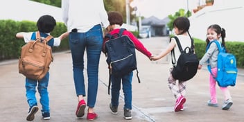 Mom and three children holding hands, wearing backpacks and walking together.