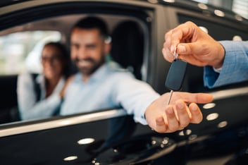 Close-up of a person's hand receiving car keys from a dealer, with a happy couple smiling inside the new car, illustrating the benefits of auto loan pre-approval.
