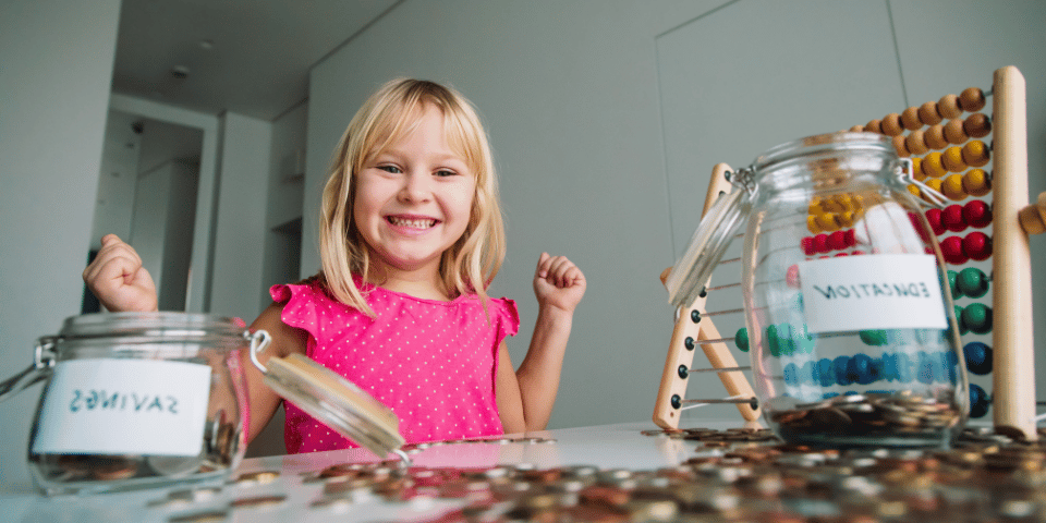 Little girl smiling in front of her coins and glass jars for savings and education.