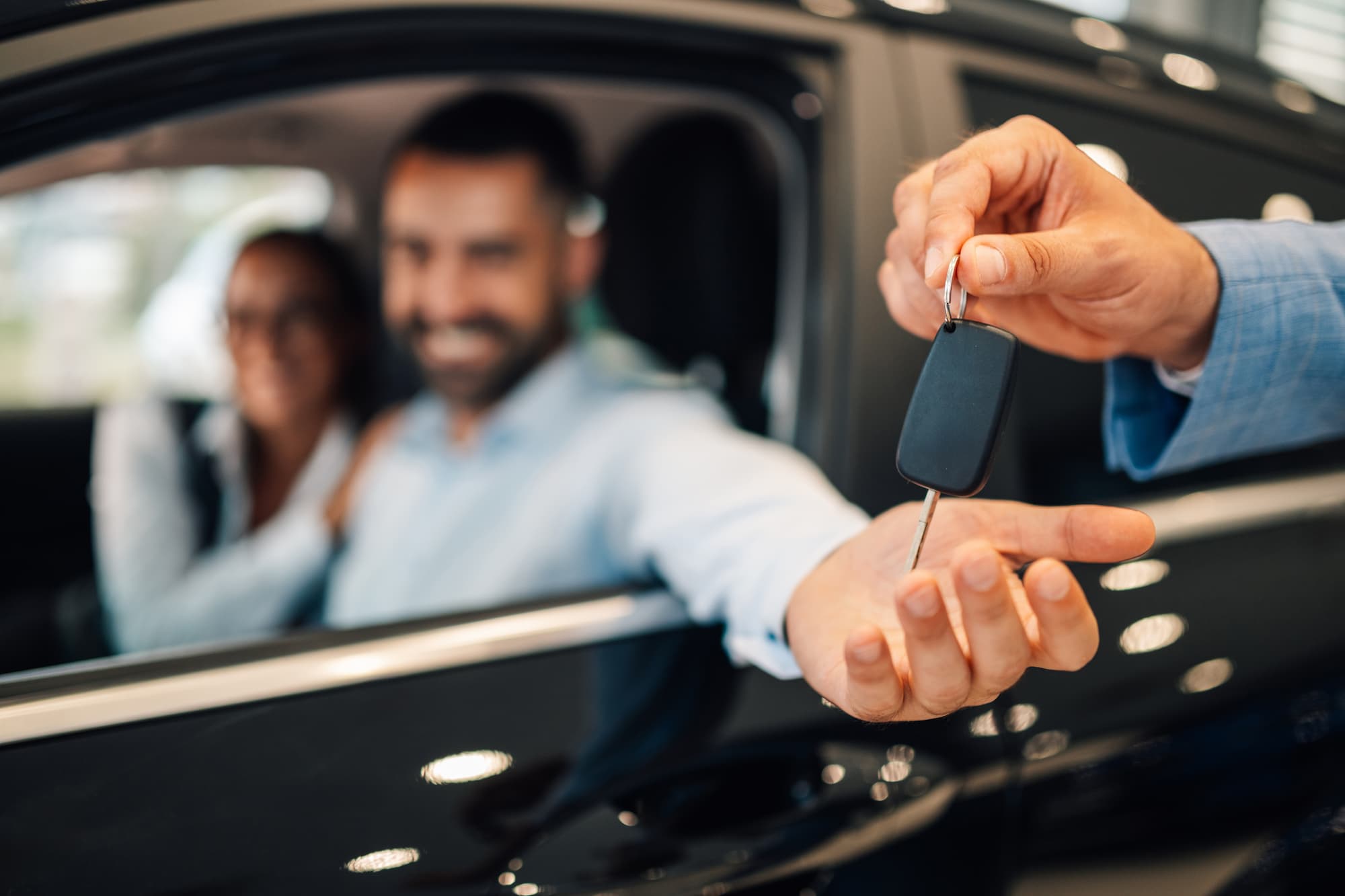 Close-up of a person's hand receiving car keys from a dealer, with a happy couple smiling inside the new car, illustrating the benefits of auto loan pre-approval.