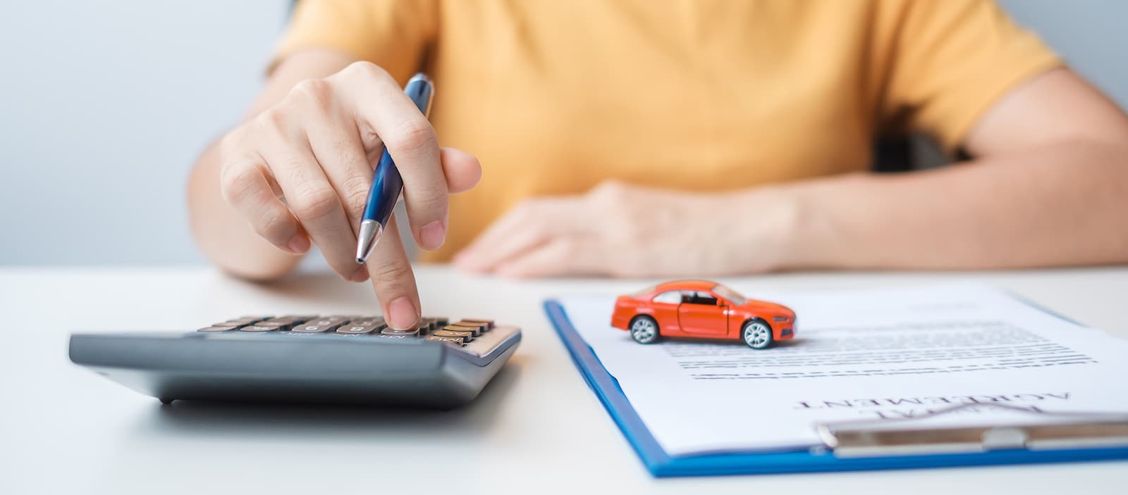 A close-up of a person using a calculator and holding a pen next to a small toy car resting on a loan agreement document.